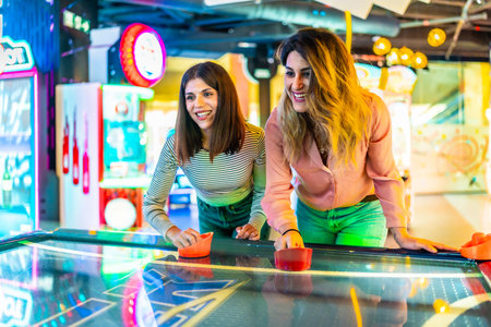 Two young women enjoying a friendly game of air hockey in a vibrant arcade, showcasing fun and entertainmentの写真素材