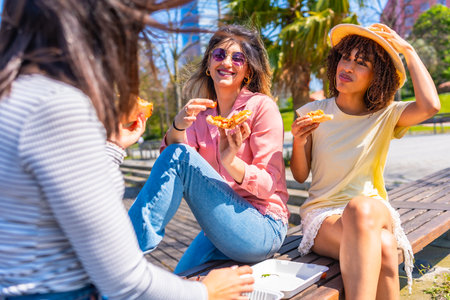 Young multi ethnic women having fun eating pizza slices in a park during a beautiful summer dayの写真素材