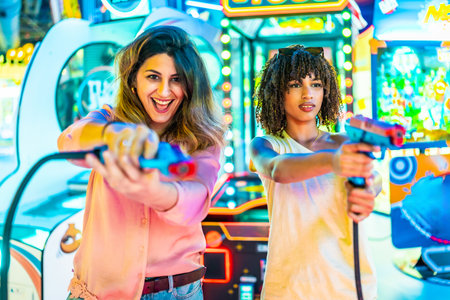 Two women enjoying their free time playing in an arcade with toy guns, laughing and having funの写真素材