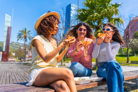 Three young women savoring delicious pizza while sitting on a bench in a vibrant city park, enjoying a sunny summer day togetherの写真素材