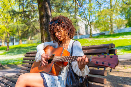 Curly haired woman sitting on a bench playing an acoustic guitar in a green city park during a sunny spring dayの写真素材