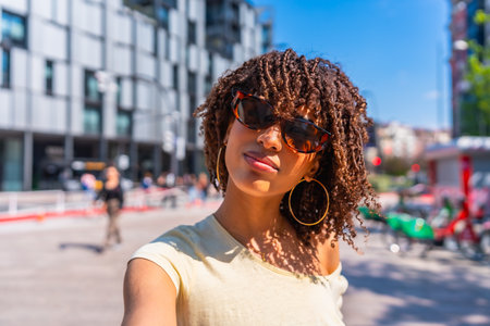 Portrait of a young woman with curly hair and sunglasses taking a selfie on a sunny day in the cityの写真素材