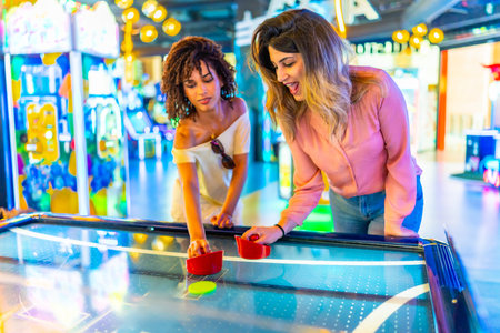 Two young women enjoying a friendly game of air hockey in a vibrant arcade, showcasing the fun and excitement of indoor entertainmentの写真素材