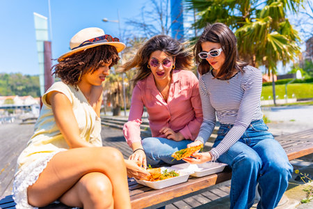Three female friends enjoying a pizza lunch together on a park bench during a sunny summer dayの写真素材