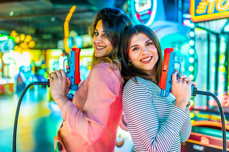 Two joyful women standing back to back, holding toy guns and enjoying playful moments in a vibrant, brightly lit amusement arcadeの写真素材