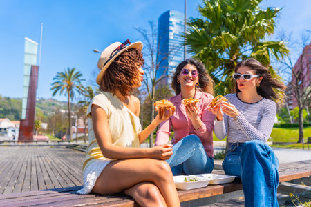 Three women savoring pizza slices while enjoying a sunny day on a park bench, sharing laughter and creating lasting memories in the cityの写真素材