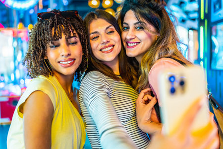 Smiling friends capturing a joyful selfie with a smartphone in a vibrant amusement arcade filled with colorful lights and excitementの写真素材