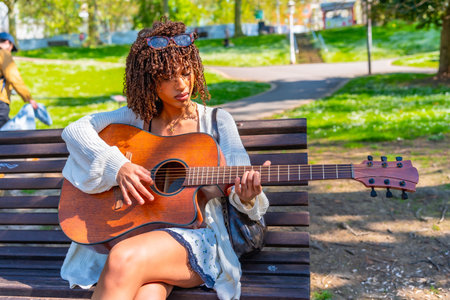 Musician concentrating while playing acoustic guitar sitting on a bench in a green city park during a sunny spring dayの写真素材