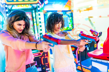 Two young women focusing intently while enjoying a thrilling shooting game in a vibrant amusement arcade filled with colorful lightsの写真素材