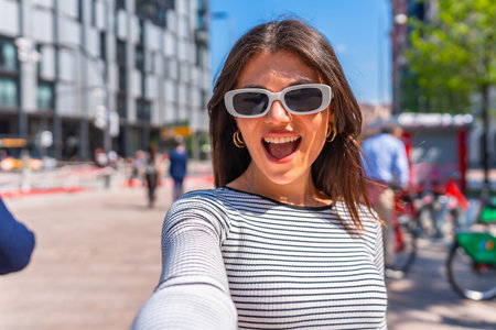 Happy woman wearing trendy sunglasses, joyfully taking a selfie in the bustling city center on a bright summer dayの写真素材