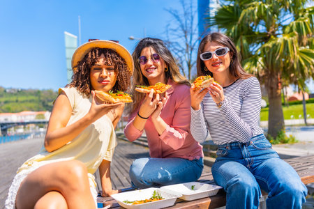 Three women enjoying pizza slices while sitting on a bench in a sunny city park, sharing laughter and savoring a delightful summer dayの写真素材