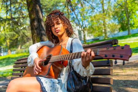 Young woman sitting on a park bench, playing an acoustic guitar while enjoying a sunny day filled with music and relaxation outdoorsの写真素材