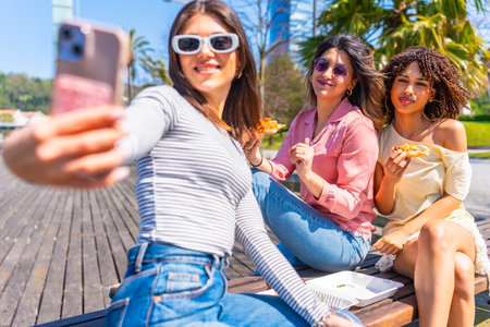 Group of female friends taking a selfie while enjoying pizza on a bench in a city park during a sunny summer dayの写真素材