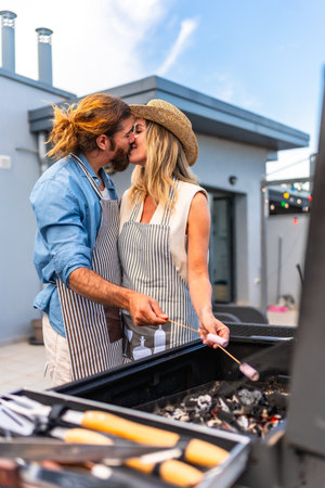 Romantic couple sharing a kiss while grilling marshmallows during a barbecue on a rooftop terraceの写真素材