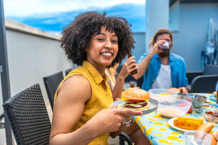 Young woman showing her delicious burger, enjoying a rooftop barbecue party with friends, drinking and having fun togetherの写真素材