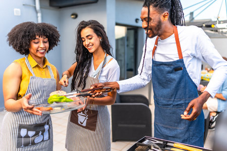 Happy multi ethnic friends preparing grilled vegetables and meat, enjoying barbecue party on the rooftopの写真素材