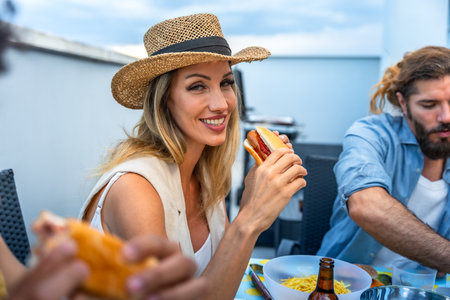 Happy woman eating hot dog at a rooftop barbecue party with friends, enjoying good food and companyの写真素材