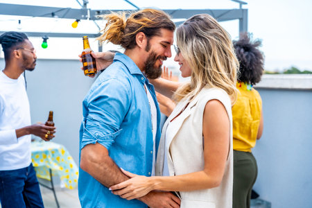 Happy couple embracing tenderly at a rooftop party, surrounded by friends enjoying drinks and good companyの写真素材