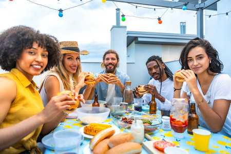 Cheerful multi ethnic friends savoring delicious burgers while enjoying a vibrant rooftop barbecue party with stunning city skyline viewsの写真素材