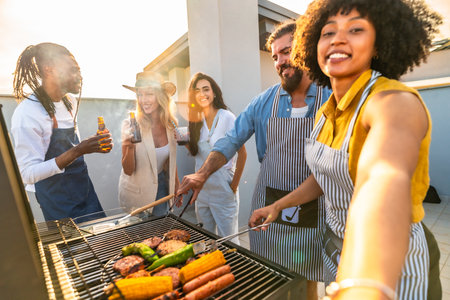 Cheerful multi ethnic friends are having fun, drinking beer and taking selfie while preparing barbecue on a rooftop at sunsetの写真素材