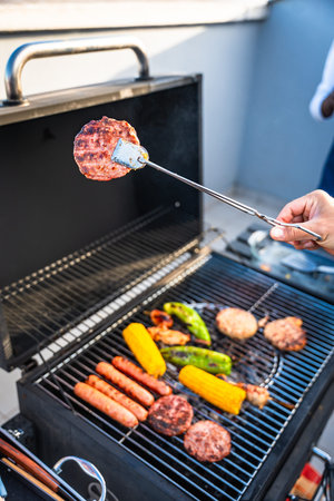 Hand using tongs to grill hamburgers, sausages, corn and peppers on a barbecue grill during a summer...