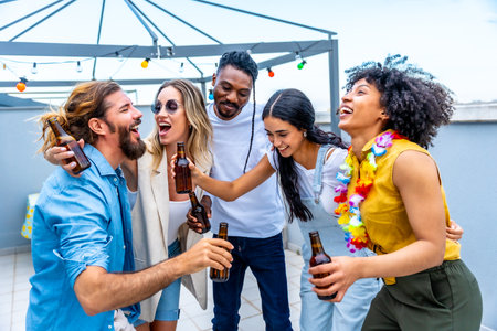 Cheerful multi ethnic friends are toasting with bottles of beer, laughing and having fun at a rooftop partyの写真素材