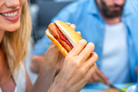 Woman enjoying a hot dog with ketchup, sitting at a picnic table, with a blurred man in the backgroundの写真素材