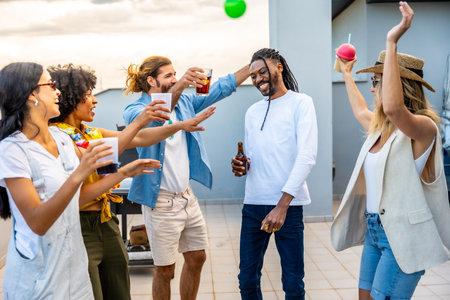 Cheerful young people enjoying drinks and dancing joyfully at a vibrant rooftop party, creating unforgettable memories under the sunsetの写真素材