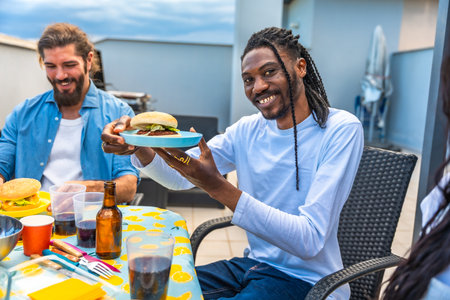 Cheerful friends enjoying a rooftop barbecue party, sharing burgers, drinks, and laughter on a sunny dayの写真素材
