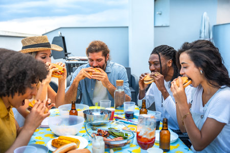 Cheerful multi ethnic friends savoring delicious burgers while enjoying a lively rooftop barbecue party, celebrating summer togetherの写真素材
