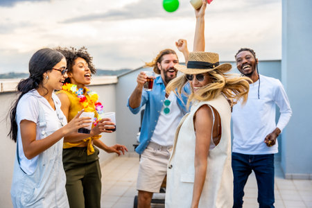 Cheerful multi ethnic friends enjoying cocktails while dancing joyfully at a vibrant rooftop party, celebrating summer under the sunsetの写真素材