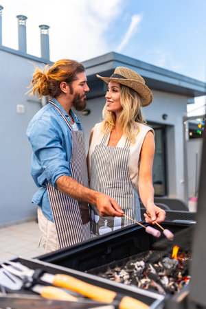 Happy couple enjoying a sunny summer day, grilling marshmallows on a rooftop barbecue, sharing laughter and creating joyful memories togetherの写真素材