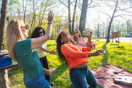 Four multi ethnic women dancing and having fun in a park, one of them is filming with her phoneの写真素材