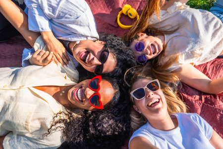 Four cheerful young women wearing sunglasses are lying together on a blanket in a park, happily laughing and enjoying their time togetherの写真素材