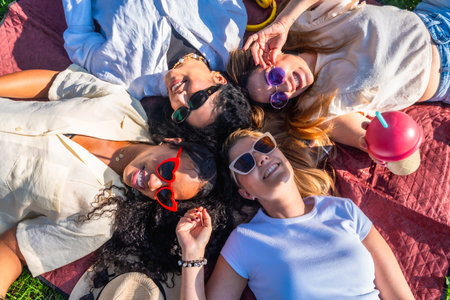 Four diverse female friends are relaxing on a blanket in a park, enjoying a sunny day togetherの写真素材