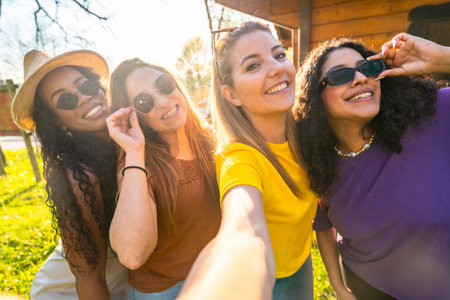 Four friends are taking a selfie outdoors, wearing sunglasses and smiling, enjoying a sunny day and having fun togetherの写真素材