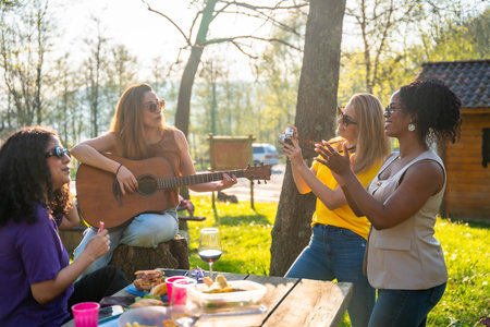 Four friends are having a picnic outdoors, with one playing guitar, one taking photos, and the others enjoying the music and foodの写真素材