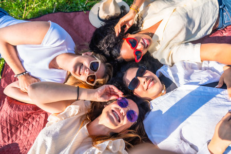 Four diverse young women lying together in a park on a blanket wearing sunglasses and smiling up at the cameraの写真素材