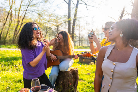 Four young women enjoying a picnic in a park, singing, playing guitar, taking pictures, and having funの写真素材