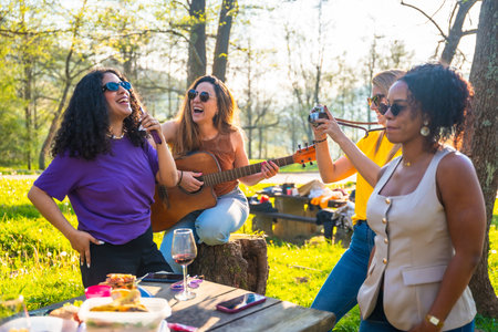 Four female friends enjoying a picnic in a park, singing, playing guitar, taking photos, and having funの写真素材