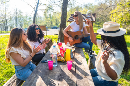 Four diverse women are having a picnic in a park, playing guitar, clapping, drinking wine, and enjoying the sunny weatherの写真素材