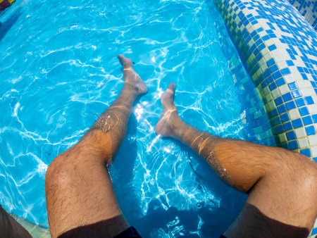 Point of view shot capturing a man relaxing in a swimming pool, legs submerged in the refreshing turquoise water under the sunny skyの写真素材