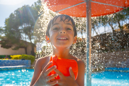 Smiling boy holding an orange water toy under a splash of water, enjoying a sunny day by the pool with trees in the backgroundの写真素材