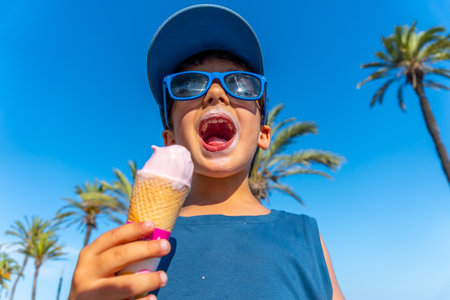 Young boy with sunglasses and cap enjoying a strawberry ice cream cone on a hot summer day with palm trees and blue skyの写真素材