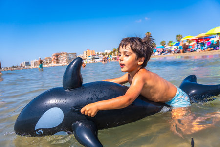 Happy child riding on an inflatable orca toy in the sea, splashing through waves and enjoying a joyful summer vacation by the beachの写真素材