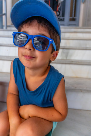 Young child sitting on steps wearing a blue baseball cap and blue sunglasses, enjoying a sunny dayの写真素材