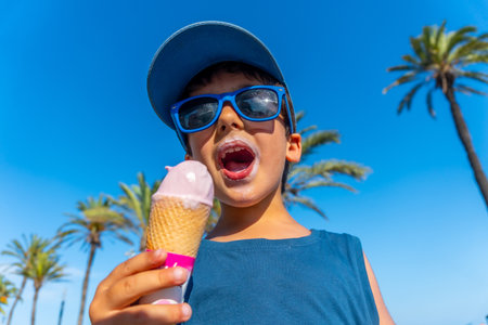 Young boy with sunglasses and blue cap enjoying a strawberry ice cream cone on a summer day with palm trees and blue skyの写真素材