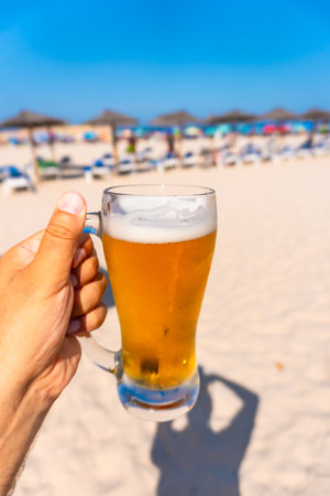 Hand holding glass of cold beer on beach with blurred beach umbrellas and sunbeds in backgroundの写真素材