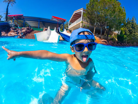 Playful child with blue sunglasses and hat pointing with finger and sticking tongue out in a swimming pool at a water parkの写真素材
