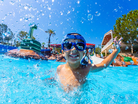 Happy boy wearing swimming goggles, splashing joyfully in a vibrant water park pool during summer vacations, enjoying every momentの写真素材